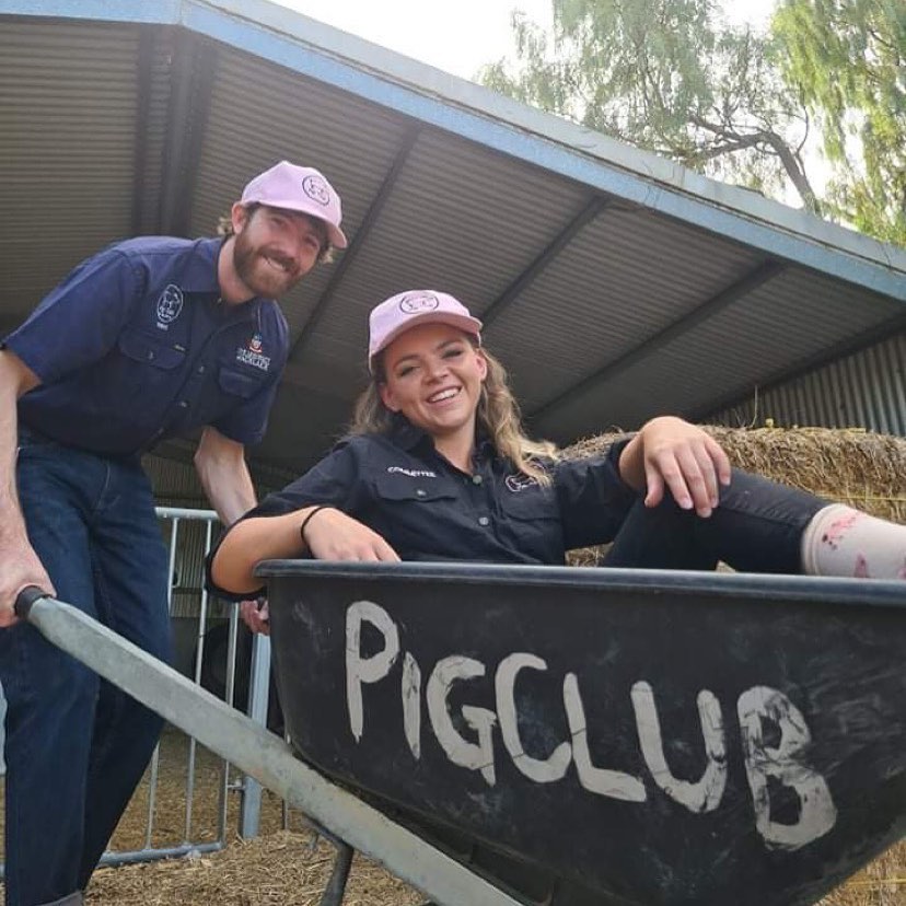 A pi club member in a pink cap pushes a wheelbarrow holding another pig club member in a pink hat