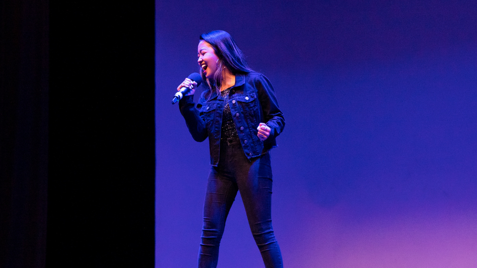 A student with long hair dressed in black denim stand on a stage and sings into a microphone under a blue light