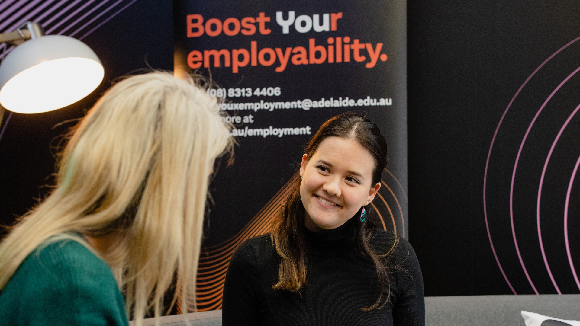 Student Employment Coordinator consults with a student holding a laptop on a grey couch.